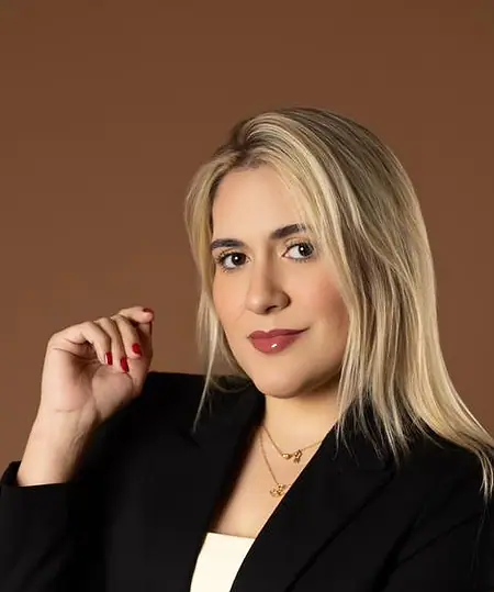 Dra. Caroline Bandeira, de blazer preto, em pé, com a mão próxima ao queixo, diante de um fundo terra