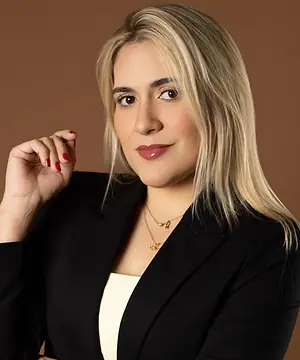 Dra. Caroline Bandeira, de blazer preto, em pé, com a mão próxima ao queixo, diante de um fundo terra
