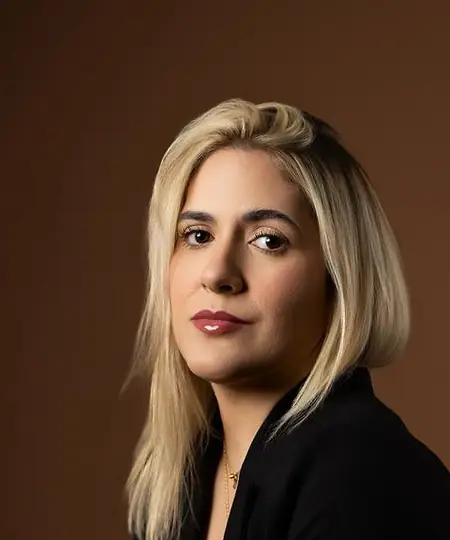 Dra. Caroline Bandeira, de blazer preto, sentada diante de um fundo terra, olhando para a câmera