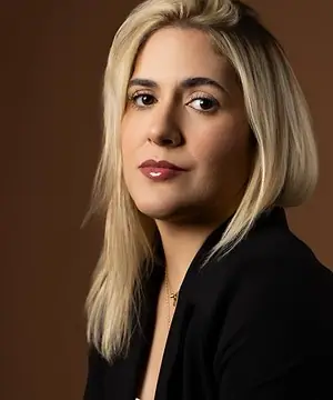 Dra. Caroline Bandeira, de blazer preto, sentada diante de um fundo terra, olhando para a câmera