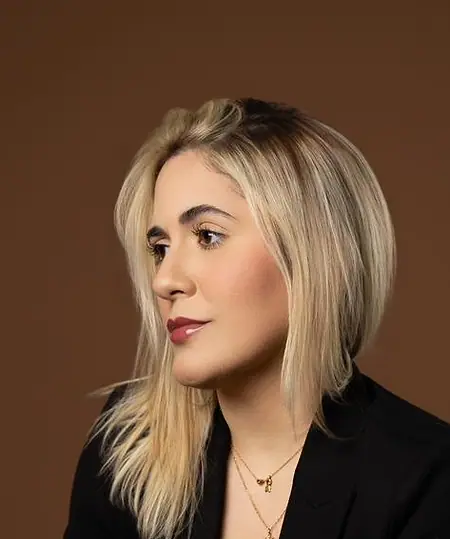 Dra. Caroline Bandeira, de blazer preto, sentada diante de um fundo terra, olhando para o lado