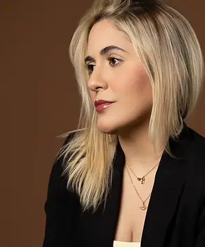 Dra. Caroline Bandeira, de blazer preto, sentada diante de um fundo terra, olhando para o lado