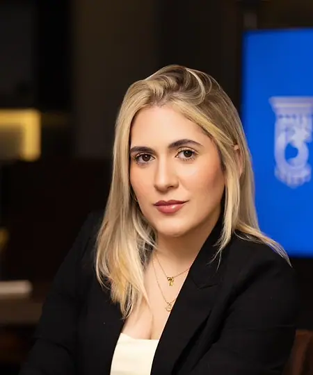 Dra. Caroline Bandeira sentada no escritório, com as mãos sobre os joelhos e o logo na tela ao fundo