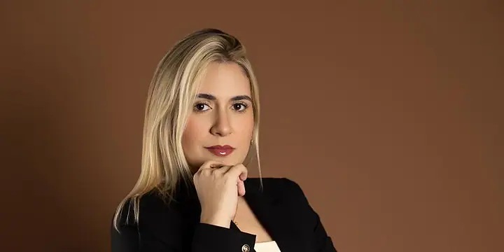Dra. Caroline Bandeira, de blazer preto, com a mão no queixo, olhando para a câmera