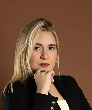 Dra. Caroline Bandeira, de blazer preto, com a mão no queixo, olhando para a câmera