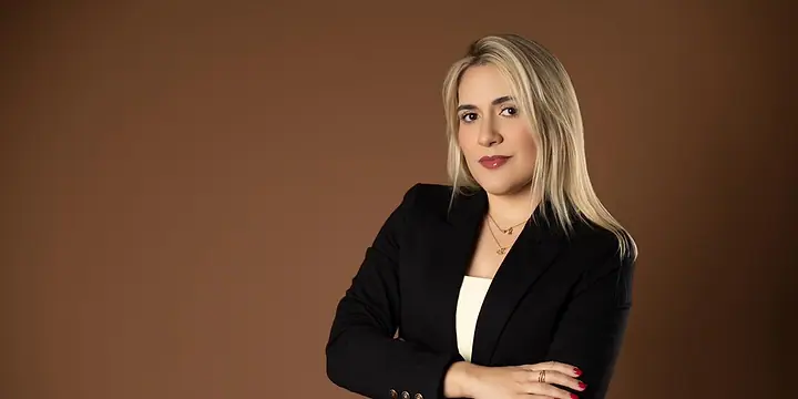 Dra. Caroline Bandeira, de blazer preto e braços cruzados, diante de um fundo terra